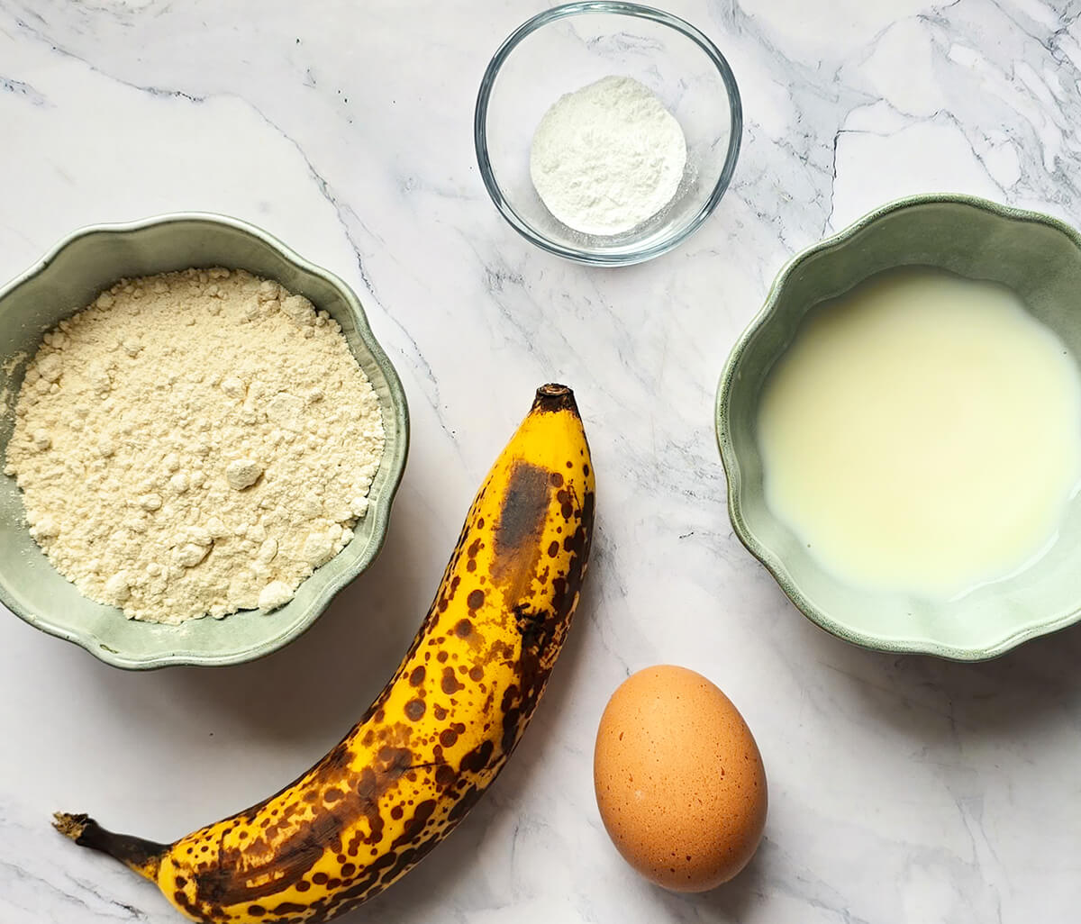 Vue de dessus d'un plan de travail en marbre blanc avec les ingrédients de la recette : une banane mûre tachetée, deux bols cannelés contenant de la farine de sarrasin et du lait d'amande, un œuf et une petite coupelle de poudre à lever.