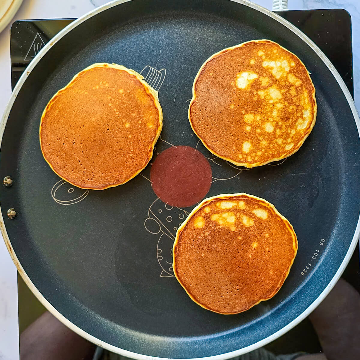 Trois pancakes sains à la banane dorés et parfaits, fraîchement cuits, sont alignés sur la poêle T-fal noire. Ils ont une texture légère et aérée, prêts à être garnis.