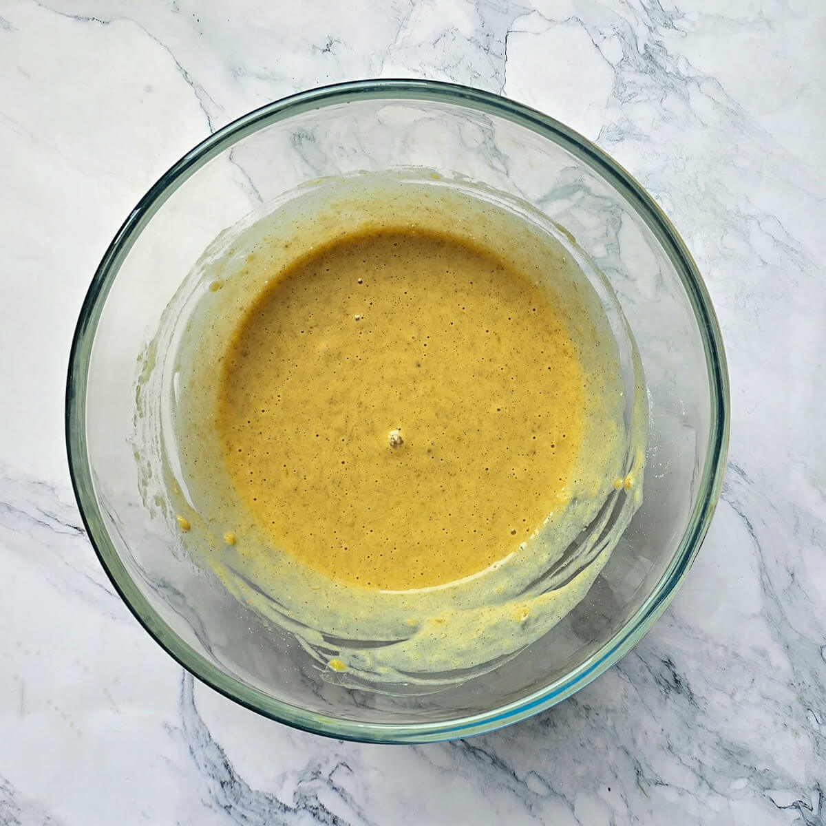 Un grand bol en verre transparent rempli de pâte à pancake à base de sarrasin et de banane écrasée, de couleur jaune d'or avec des petits grains, posé sur un plan de travail en marbre.