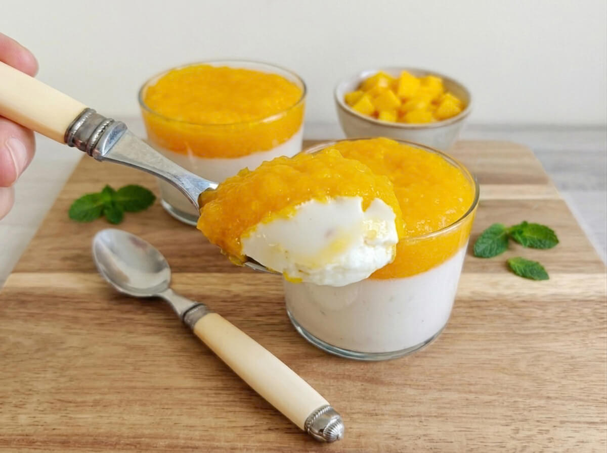 Gros plan d'une cuillère plongeant dans une verrine de panna cotta au lait de coco onctueuse, révélant la mousse blanche sous un coulis de mangue fraîche. En arrière-plan, deux verrines, un bol de mangue en dés et des feuilles de menthe sur une planche en bois.