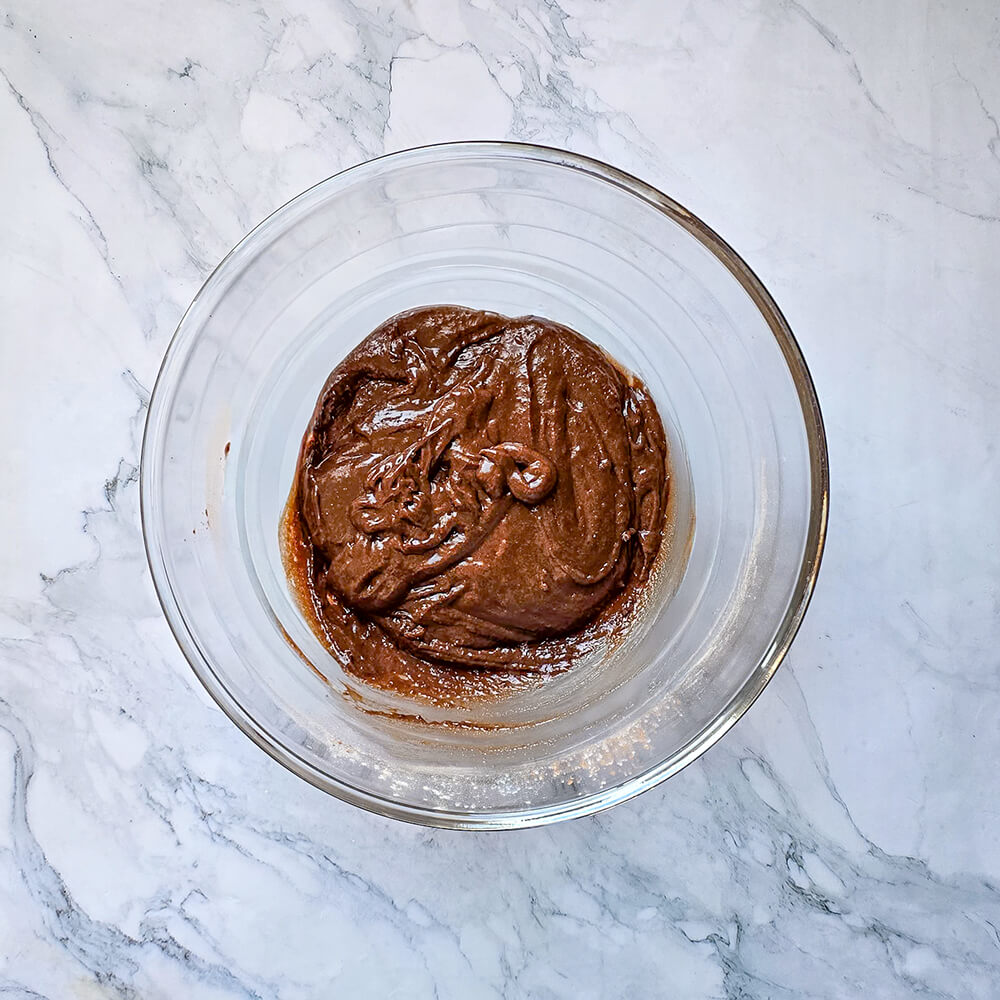 Gros plan sur une pâte à chiffon cake au cacao épaisse, onctueuse et de couleur brun foncé, fraîchement mélangée dans un saladier en verre sur un plan de travail en marbre.
