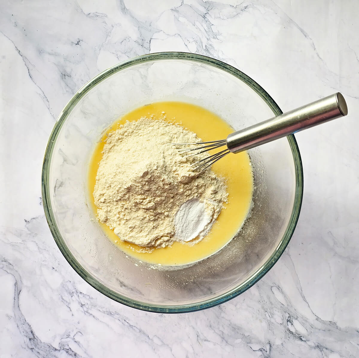 Préparation de la pâte à gâteau dans un saladier, avec un fouet, sur fond blanc marbré.