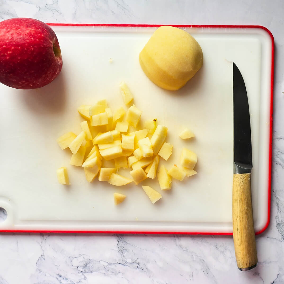 Vue de dessus d'une pomme rouge entière, d'une pomme épluchée et d'un petit tas de dés de pomme fraîche sur une planche à découper blanche avec un couteau à manche en bois.