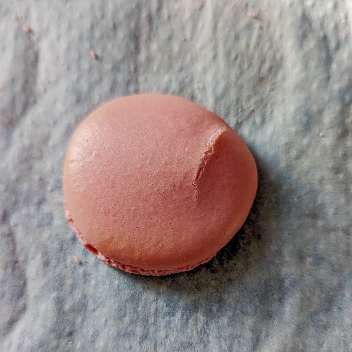 Gros plan d'une coque de macaron rose présentant une fissure nette sur le dessus, illustrant un problème de cuisson ou de croûtage sur un tapis de cuisson.