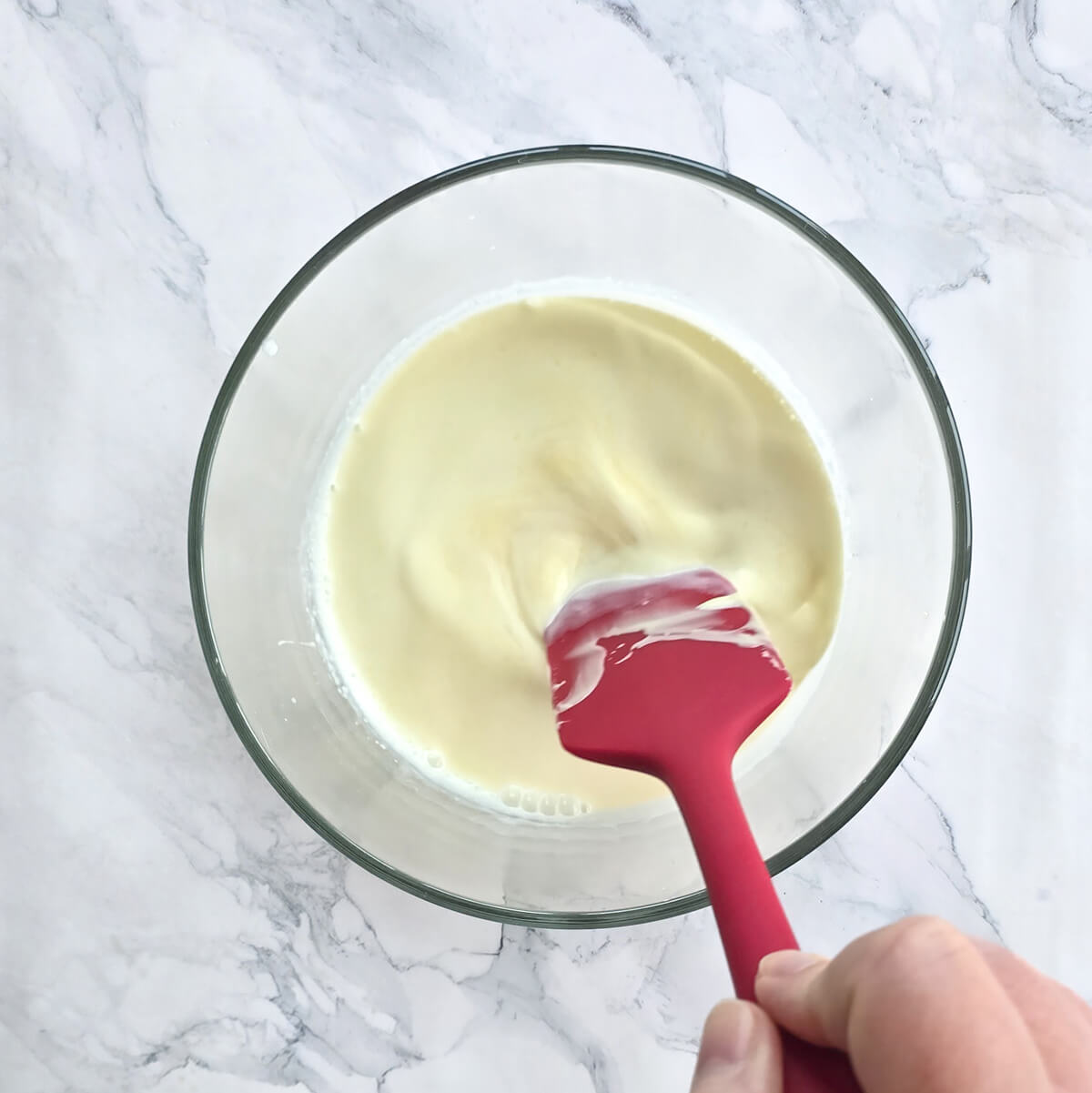 Gros plan d'une main mélangeant une préparation de ganache au citron lisse et crémeuse avec une spatule en silicone rouge dans un bol en verre.