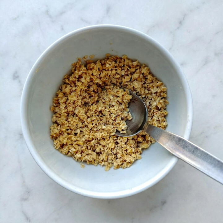 Un bol blanc contenant un mélange de flocons d'avoine et poudre d'amandes et une cuillère en métal sur un plan de travail en marbre.