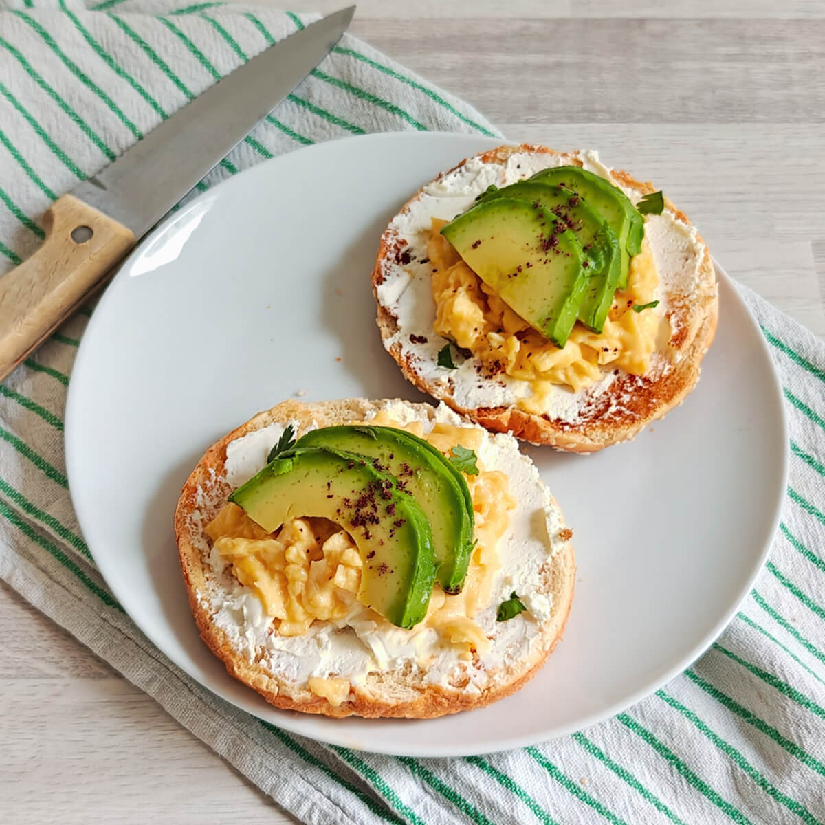 Un bagel ouvert en deux, œufs brouillés et avocat en lamelles sur le dessus, sur une assiette blanche, couteau sur le côté.