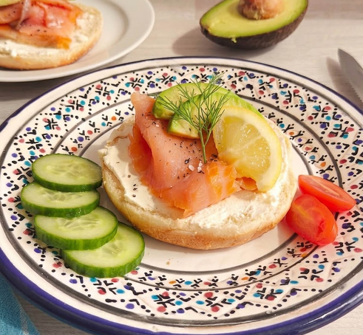 Un bagel ouvert en deux avec du fromage à tartiner, des tranches de saumon fumé, des rondelles de concombre, et des tomates cerises, sur une assiette bleue colorée.