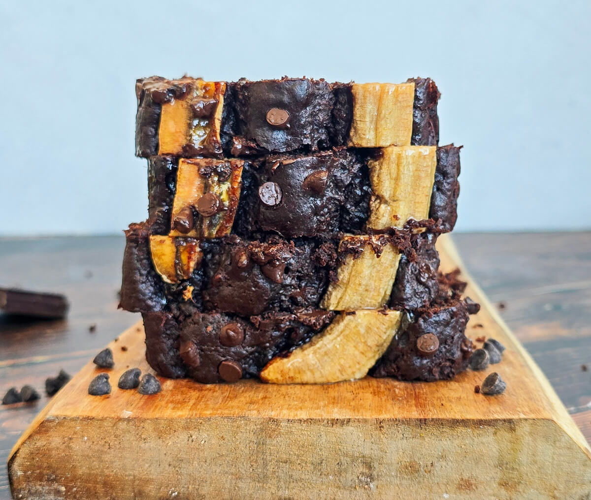 Une pile de trois tranches de banana bread au chocolat avec des morceaux de banane visible sur le dessus et parsemée de pépites de chocolat, posée sur une planche à découper en bois.