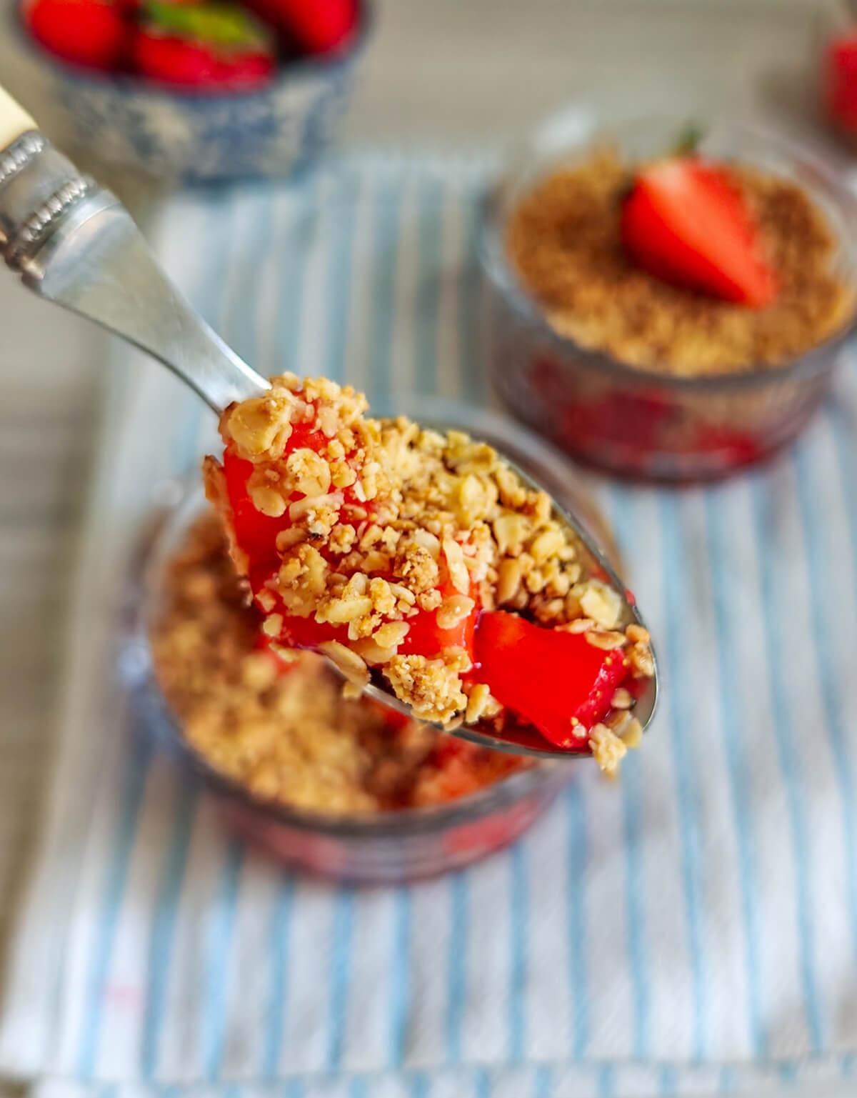 Gros plan sur une cuillère contenant une bouchée de fraises fraîches et de crumble croustillant, avec les verrines en arrière-plan flou.