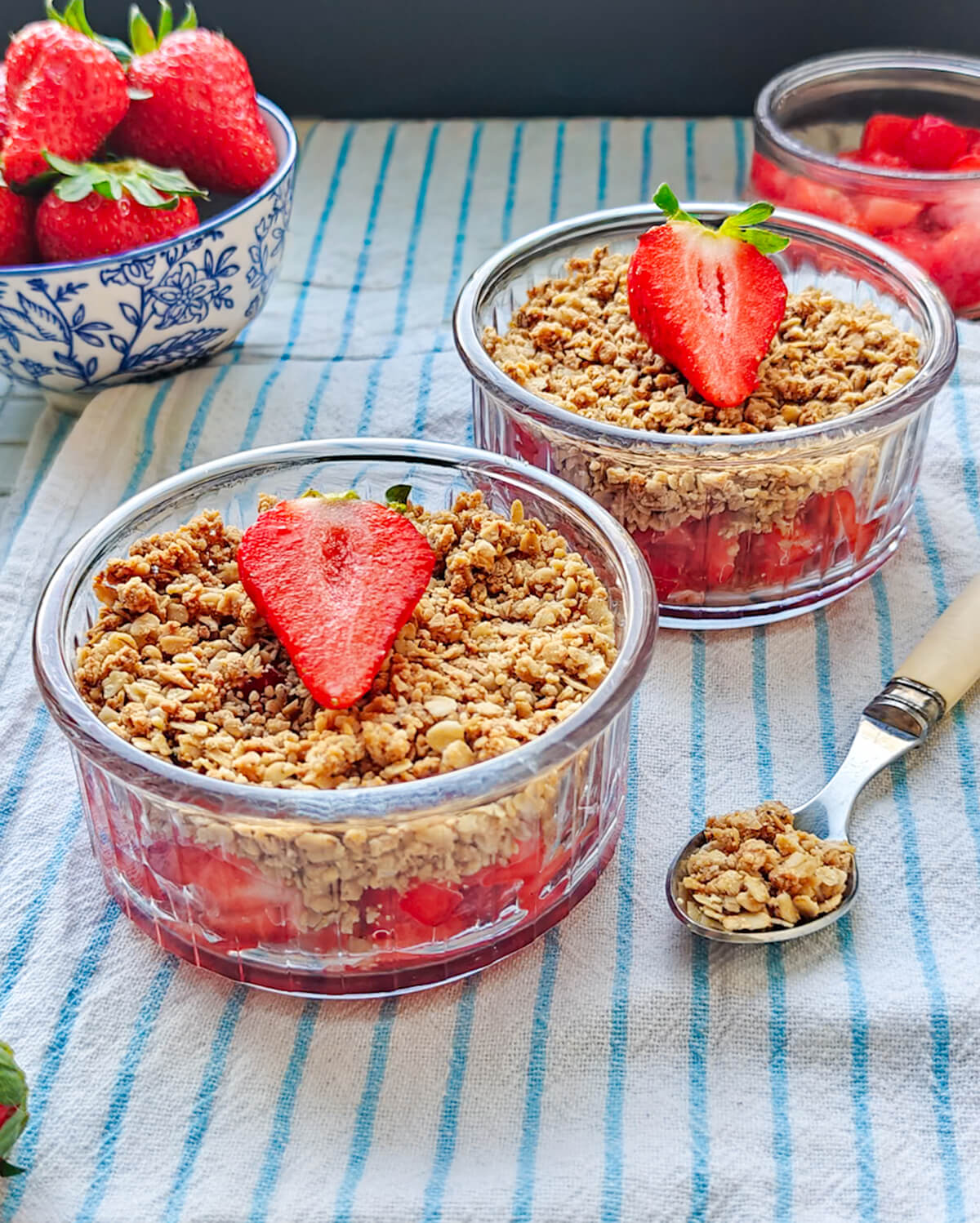 Deux verrines de crumble aux fraises décorées d'une demi-fraise, posées sur une nappe rayée à côté d'un bol de fruits frais.