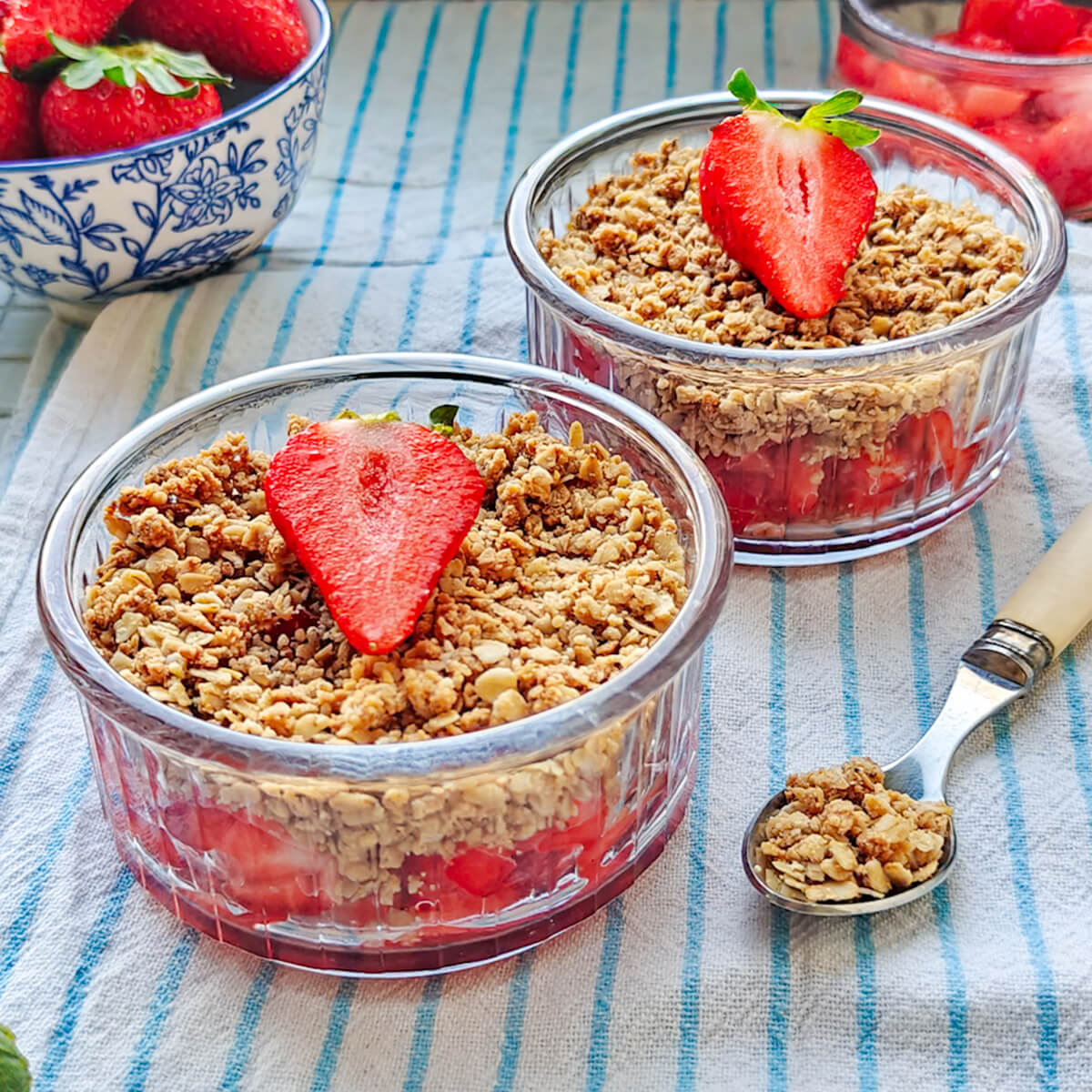 Deux verrines de crumble aux fraises décorées d'une demi-fraise, posées sur une nappe rayée à côté d'un bol de fruits frais.