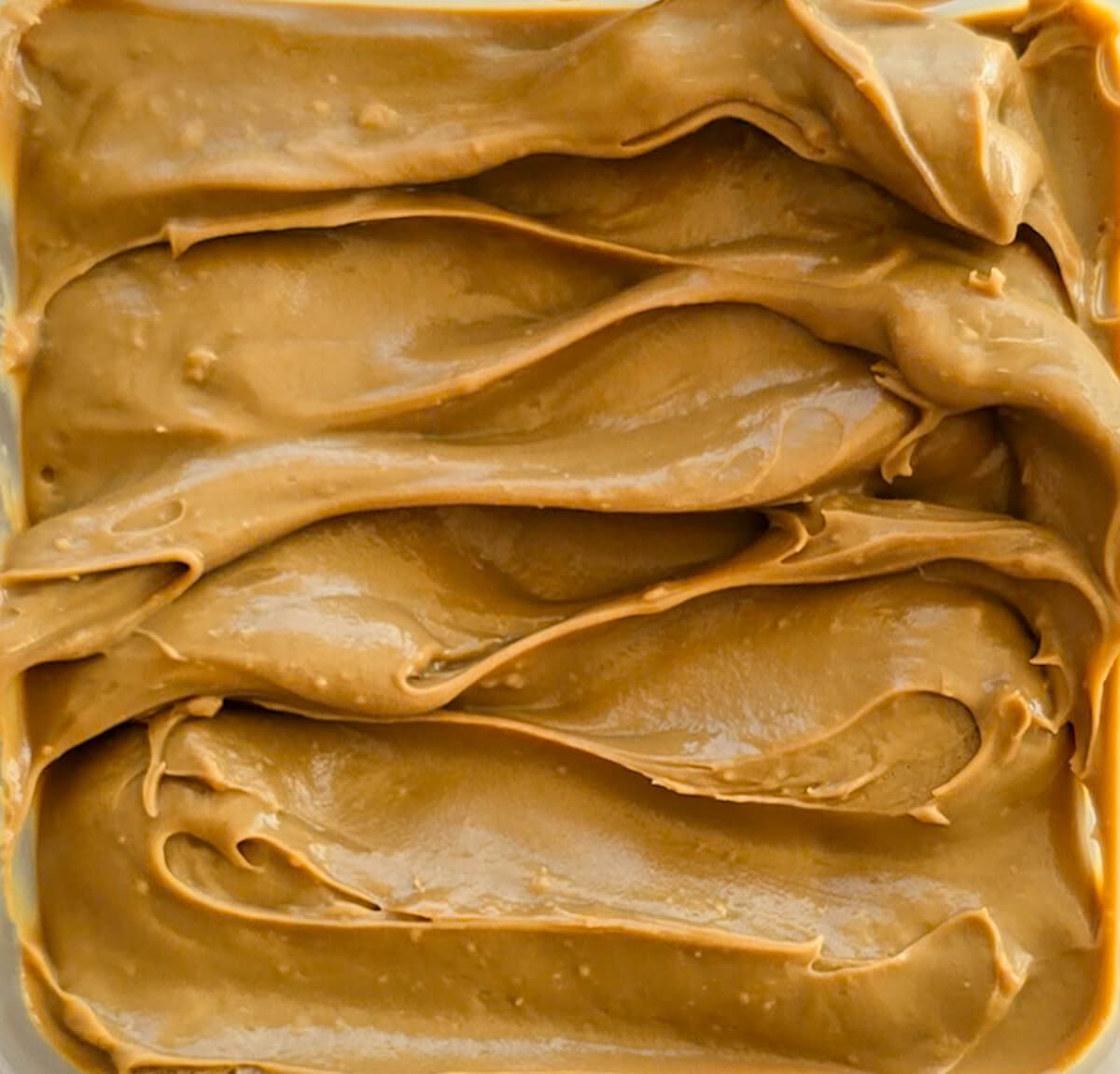 La surface d'une ganache au café dans un plat. La texture est riche, lisse et brillante, avec des ondulations profondes et des marques de spatule qui créent des motifs ondulés.