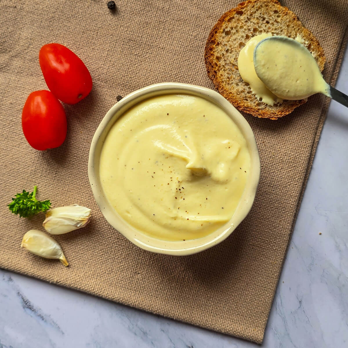 Vue de dessus d'un bol de mayonnaise œuf dur skyr sur un set de table en toile, entouré de deux tomates cerises, de gousses d'ail frais, d'un toast et d'une cuillère, sur un plan de travail en marbre.