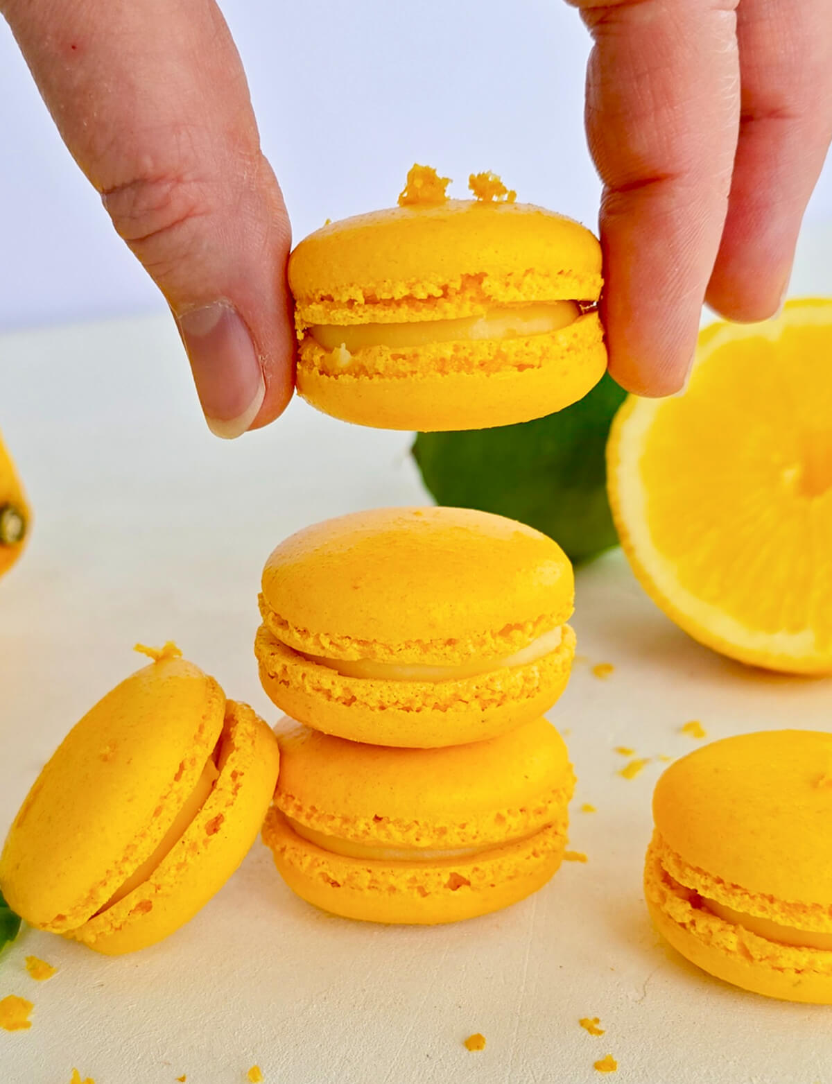 une main tenant un macaron au citron jaune, disposés de manière esthétique avec des citrons entiers et entamés en arrière-plan, parsemés de zestes frais.