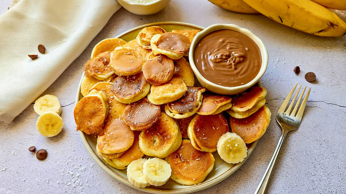 Une assiette remplie de "pancake cereal" (mini-billes de pancakes healthy à la banane) dorés et moelleux, parsemés de noix de coco râpée. Au centre, un bol de pâte à tartiner au chocolat pour le dip, avec des rondelles de banane fraîche et des pépites de chocolat en garniture.
