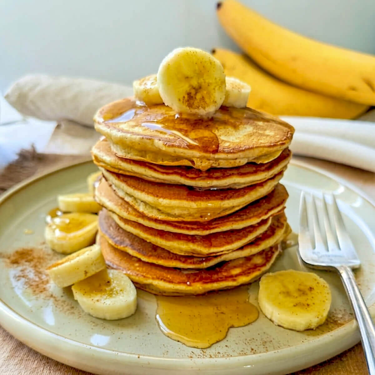 Une pile de pancakes healthy à la banane et sirop d'érable, parsemée de rondelles de banane fraîche, de cannelle servie sur une assiette en grès avec une fourchette.