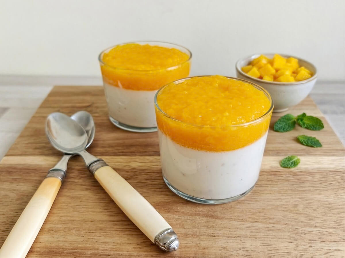 Deux verrines de dessert coco-mangue présentées côte à côte sur une planche en bois. Chaque verre montre des couches distinctes : une base de crème de coco blanche et un topping généreux de coulis de mangue jaune vif. À gauche, deux cuillères au manche ivoire et à droite, des feuilles de menthe fraîche et un bol de dés de mangue.