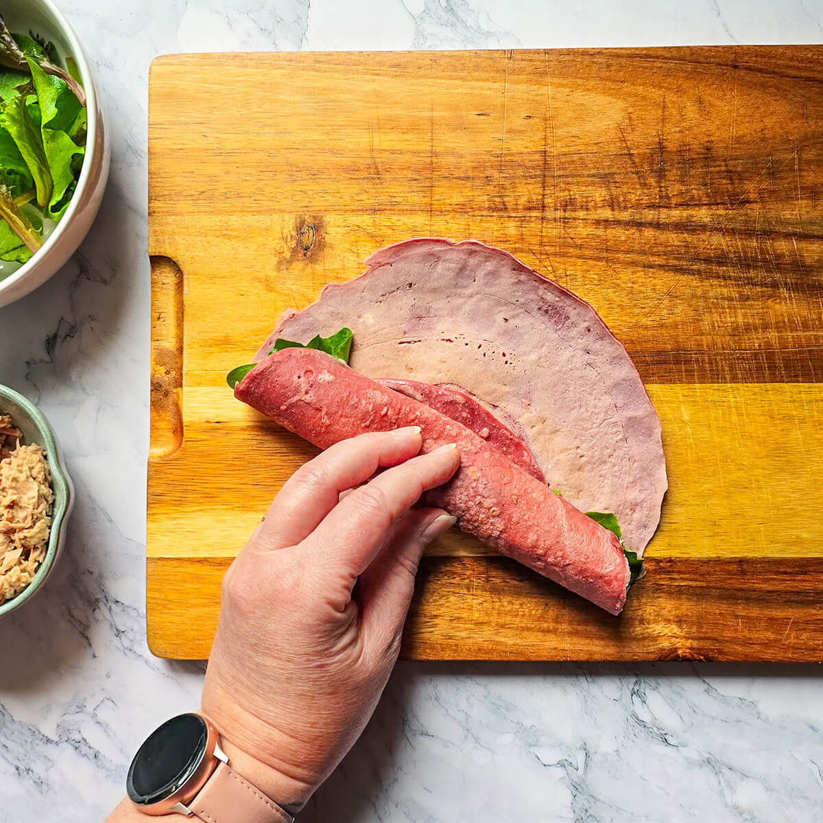 Vue de dessus montrant une main avec une montre connectée rose en train de rouler serré une galette de wrap à la betterave garnie, sur une planche à découper en bois. Le geste montre comment fermer le wrap pour préparer les bouchées apéritives. Des bols de salade et de thon sont visibles sur le plan de travail en marbre.