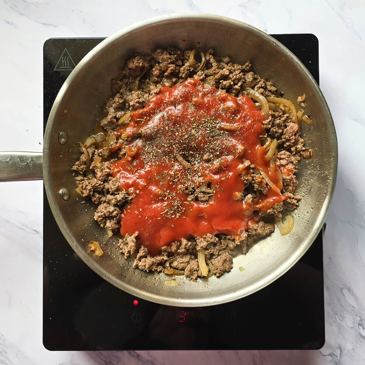 Ajout de la pulpe de tomate et des herbes de Provence sur la viande hachée cuite dans une poêle.