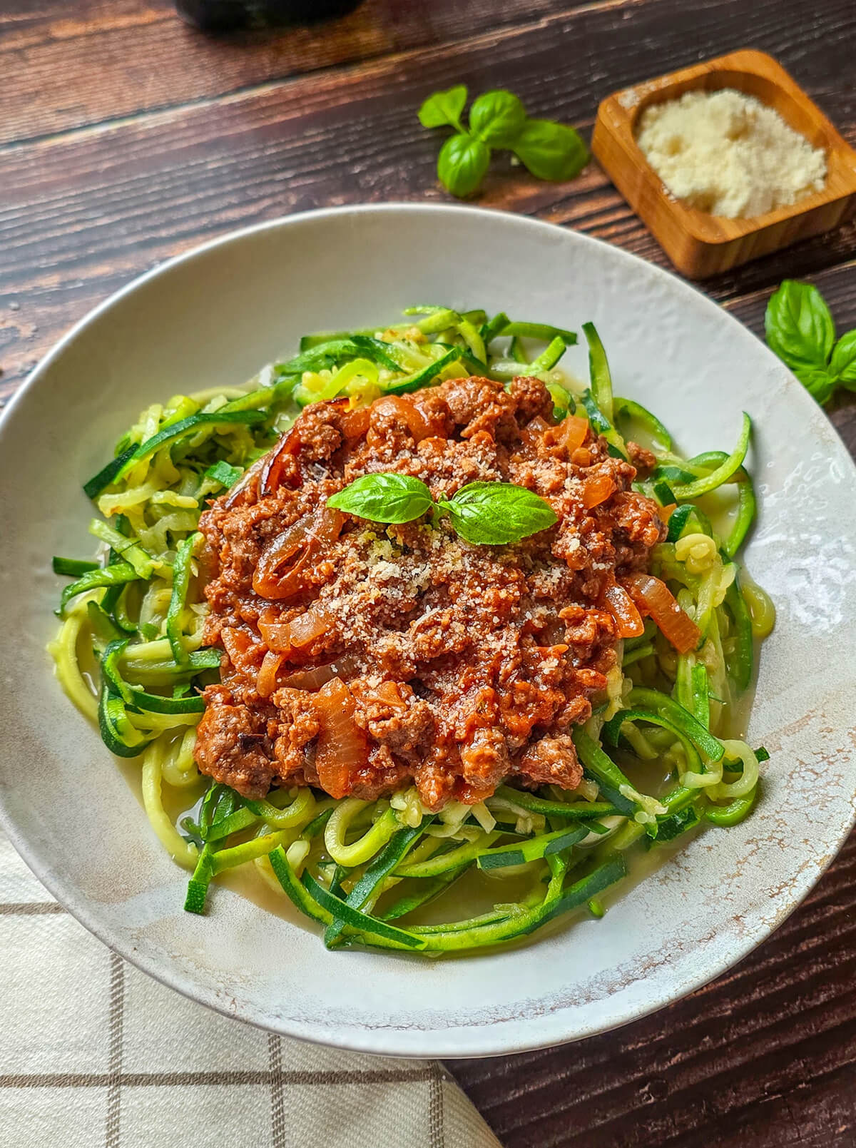 Vue aérienne (flat lay) d'une assiette de zoodles à la bolognaise. Le plat est entouré d'un torchon vichy, de feuilles de basilic frais et d'un petit bol de parmesan râpé.