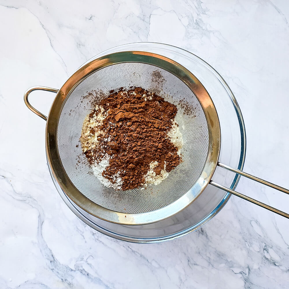 Tamis métallique posé sur un grand saladier en verre, contenant de la farine blanche et une montagne de cacao en poudre en cours de tamisage pour aérer la pâte du chiffon cake.