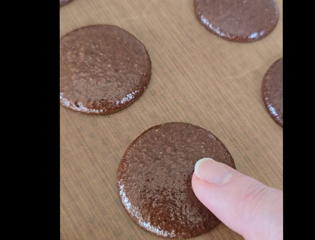 Un doigt effleurant doucement le dessus d'une coque de macaron au chocolat pour vérifier le croutage avant la cuisson, montrant une surface mate qui ne colle plus à la peau.
