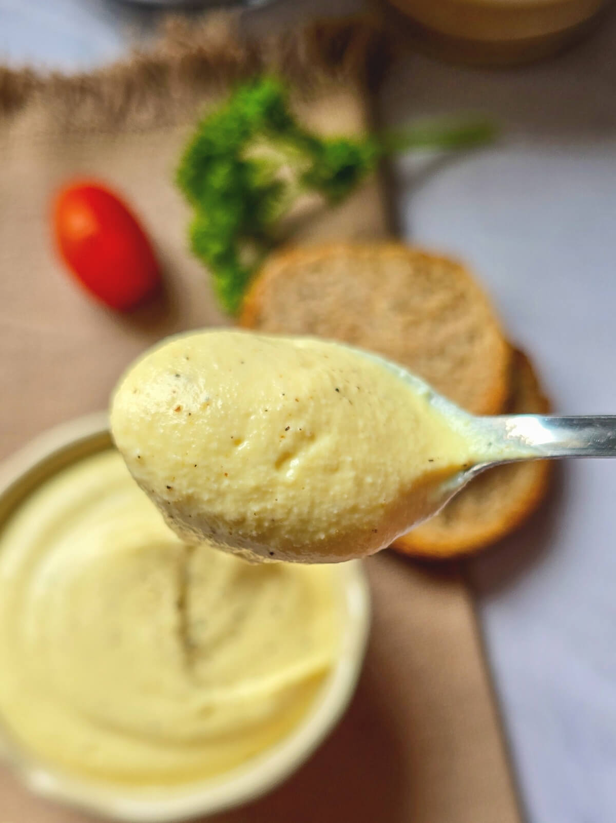 Gros plan lumineux sur une cuillère métallique remplie de mayonnaise œuf dur skyr à la texture aérienne et onctueuse, parsemée de poivre moulu, avec des toasts flous en arrière-plan.
