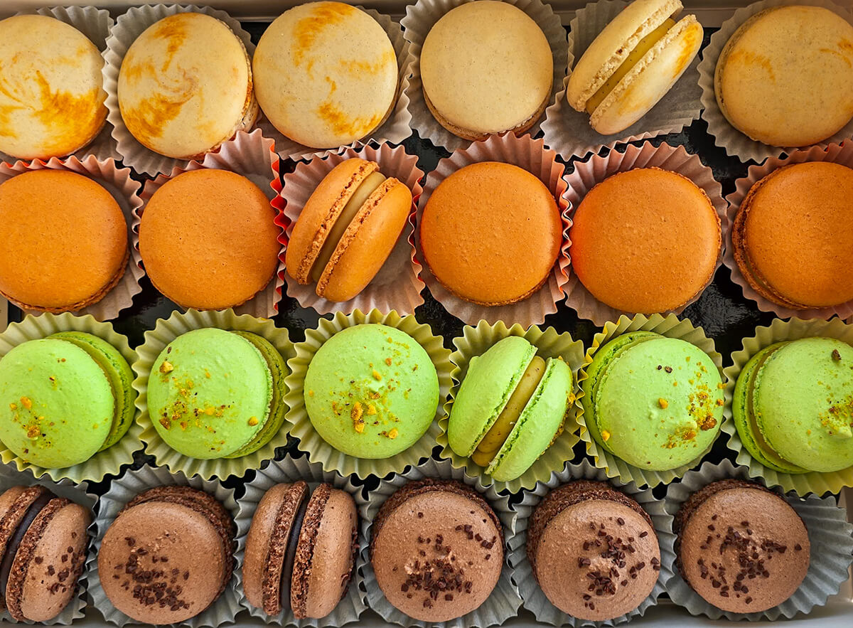 Une boîte ouverte remplie de macarons artisanaux organisés par rangées de couleurs distinctes (jaune-orange, vert-menthe, et marron-chocolat), illustrant la diversité des gammes chromatiques obtenues avec différents colorants pour macarons.
