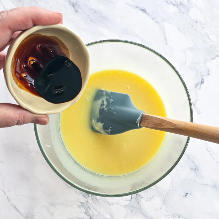 Une main versant un café serré dans un grand bol en verre contenant une préparation liquide jaune clair. Une spatule en silicone grise est déjà dans le bol en verre.