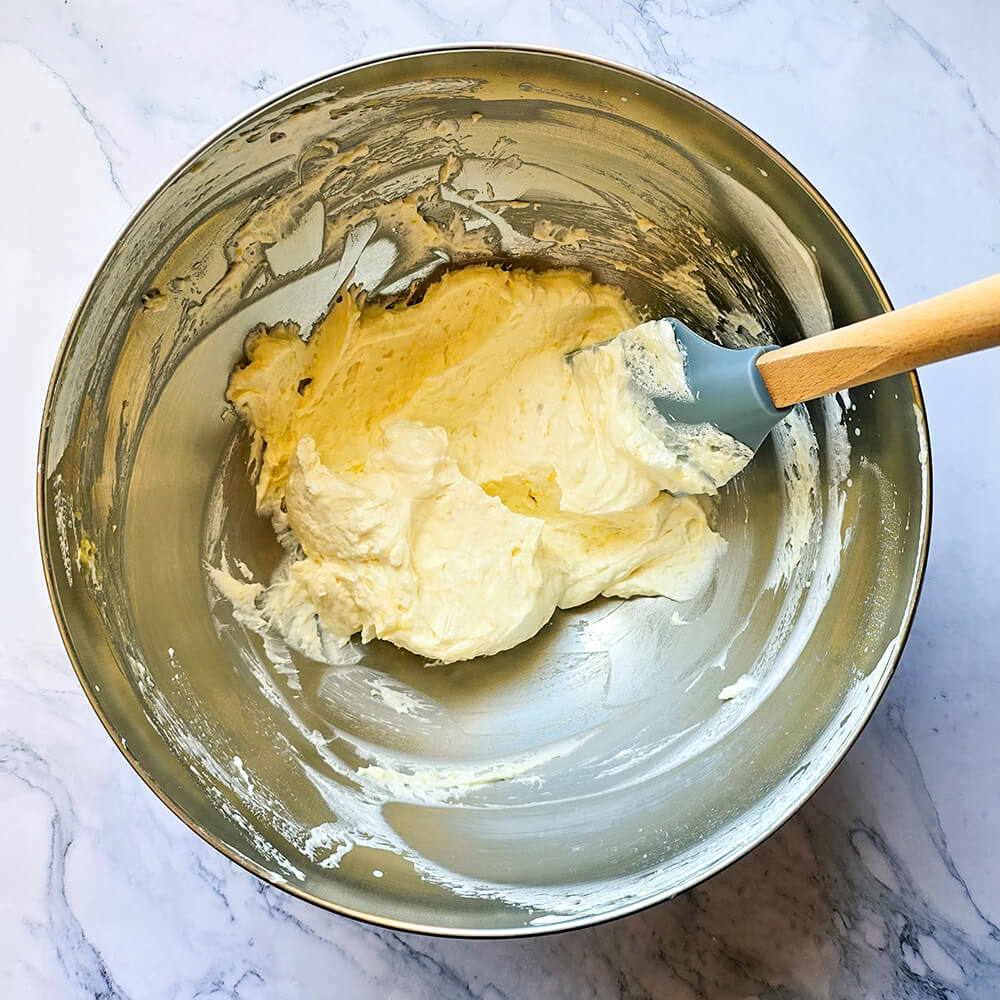 un bol en acier inoxydable contenant un mélange de crème fouettée blanche et de lemon curd jaune, créant une texture crémeuse et jaune clair. Une spatule en silicone bleue et un manche en bois sont partiellement immergés dans le bol.