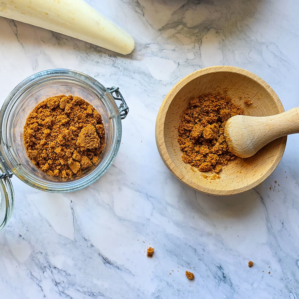 deux bols: un bocal en verre ouvert avec de la chapelure de speculoos et un bol en bois avec plus de chapelure et un pilon en bois posé à côté.