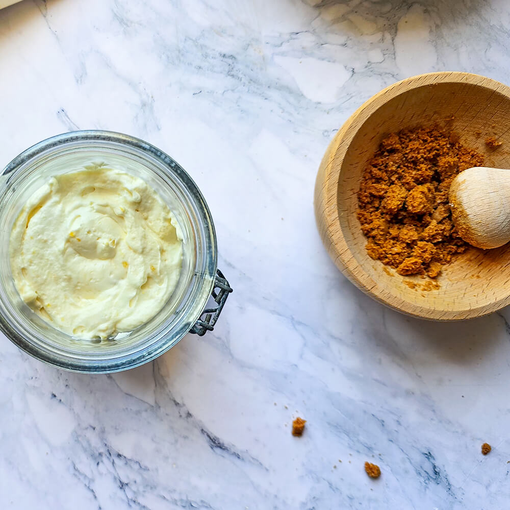 un bocal en verre ouvert contenant une couche de crème au citron et une couche de chapelure de speculoos, prête à être assemblée en verrine. Le bol en bois avec de la chapelure et le pilon sont à côté.