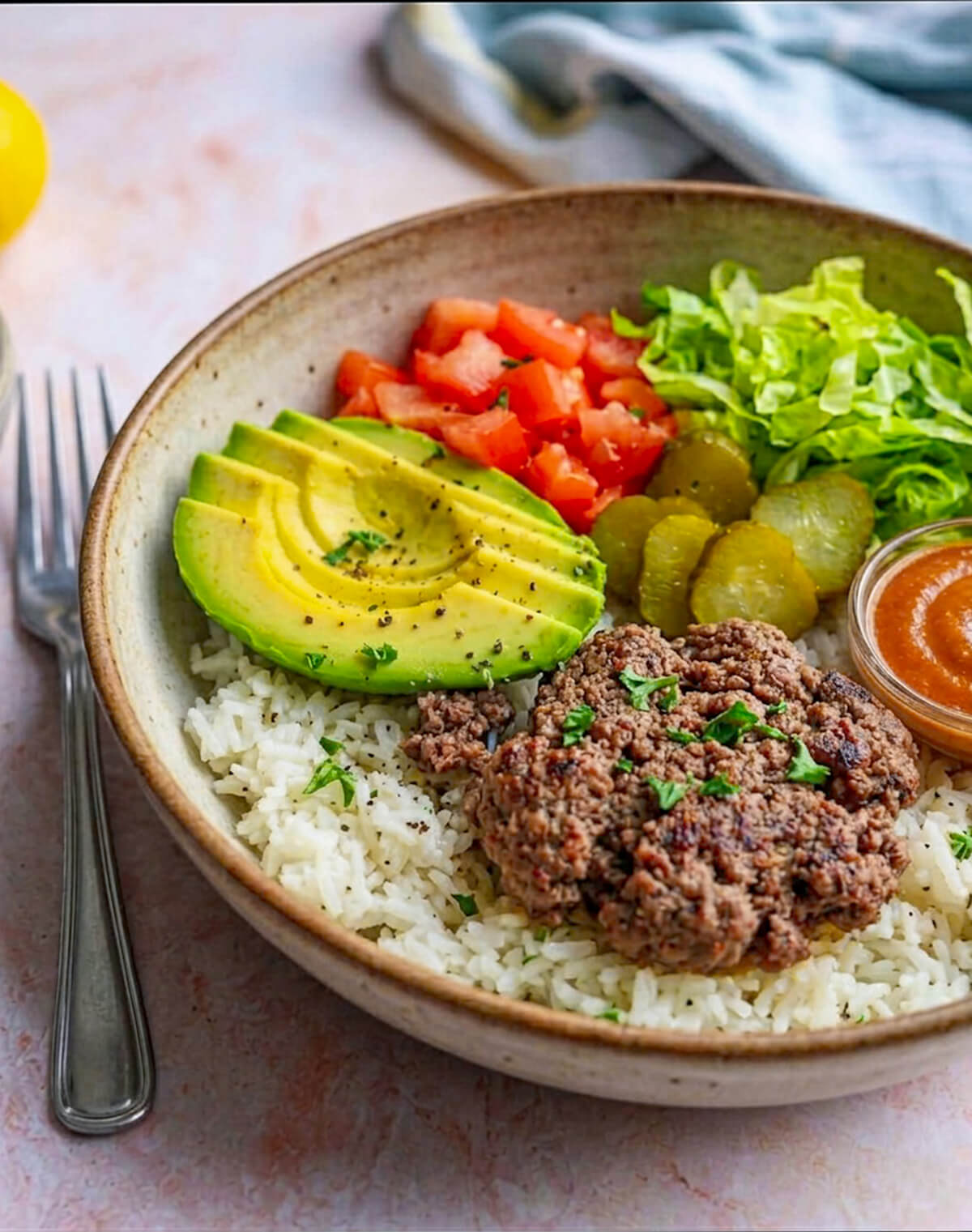 un bol beige de steak haché façon hamburger, sans pain, avec riz, tomate, avocat et salade verte sur une surface rose claire.