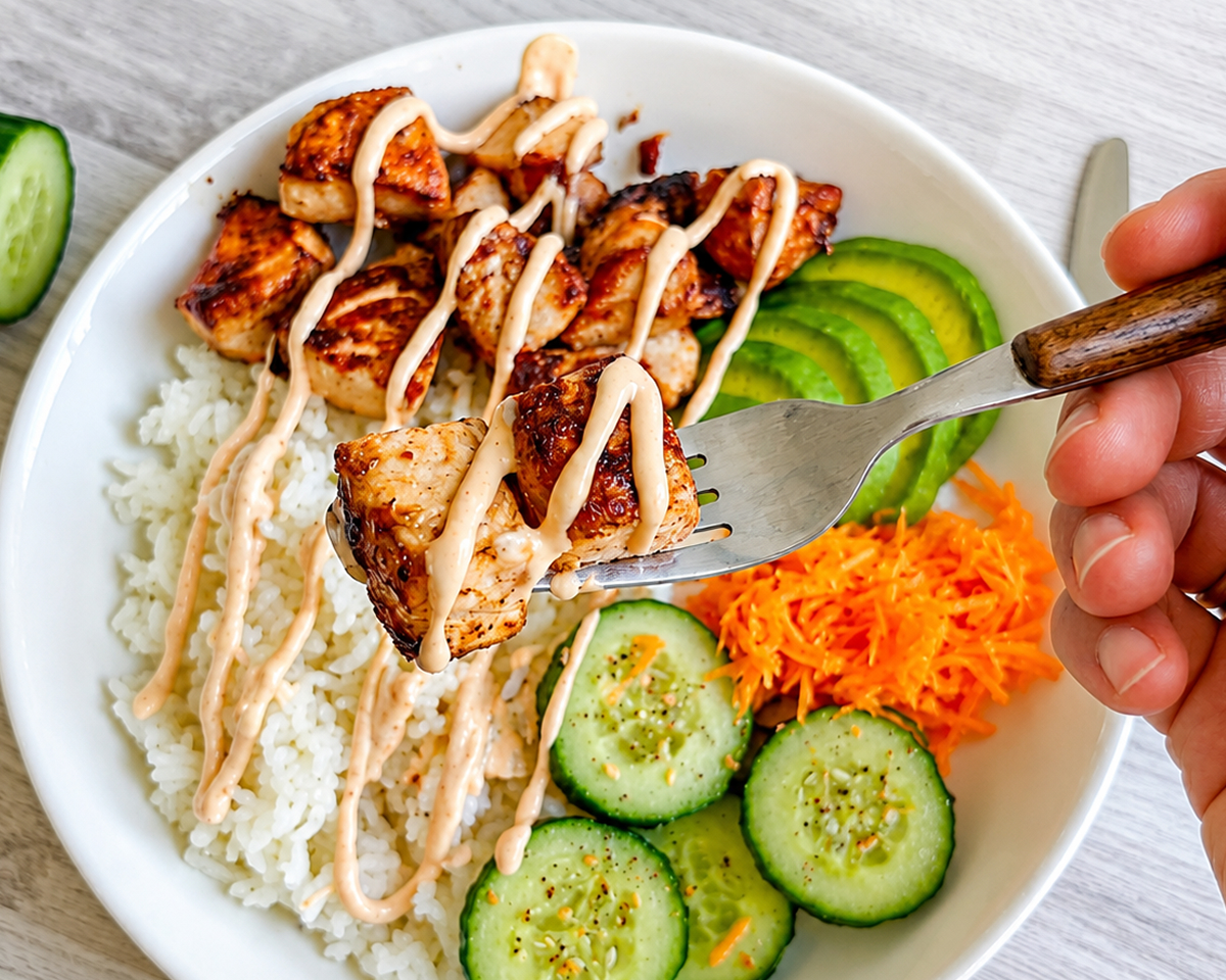 Une main tenant une fourchette avec un morceau de saumon cuit, en arrière une assiette creuse de poké bowl.