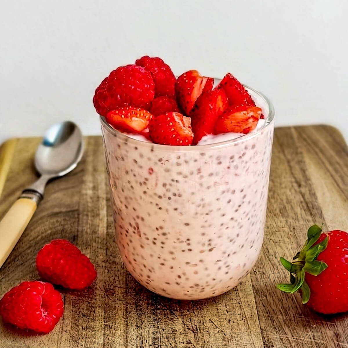 Pudding de chia à la fraise et framboise servi dans un verre transparent sur une planche à découper. Le verre est généreusement garni de fruits rouges frais. Une cuillère et quelques fruits entiers sont disposés en arrière-plan sur le bois rustique.