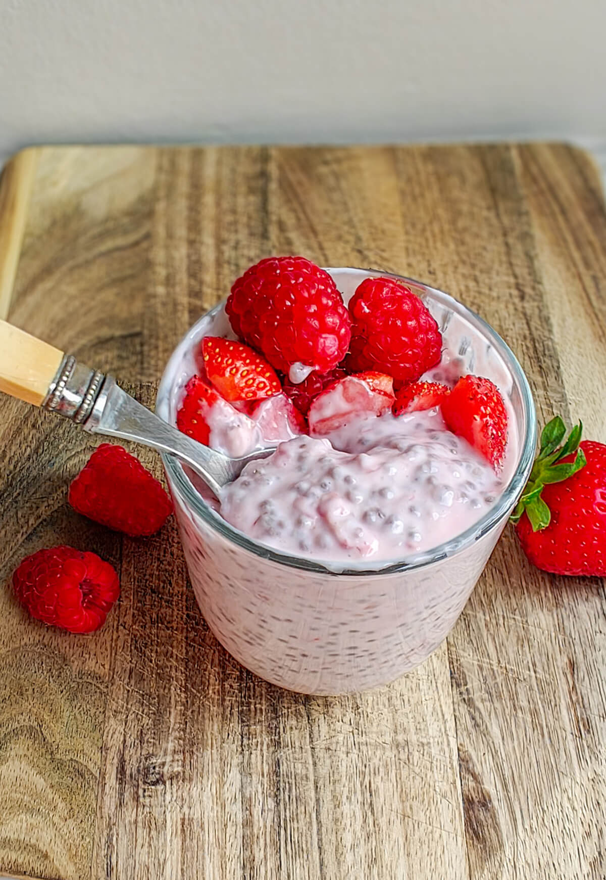 Vue rapprochée d'un pudding de chia crémeux aux fruits rouges dans un verre, posé sur une planche en bois. Une cuillère à manche clair prélève une bouchée du mélange parsemé de graines de chia. Le tout est surmonté de framboises entières et de morceaux de fraises juteuses.