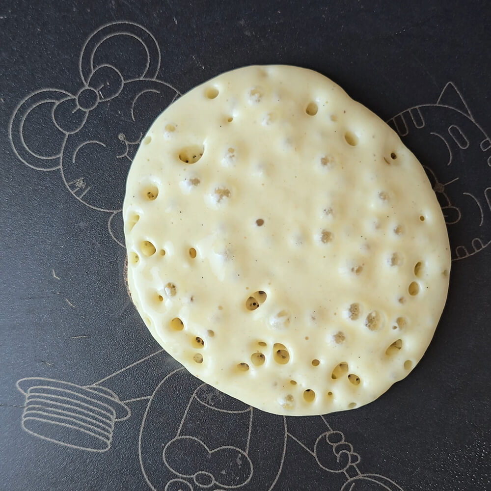 Gros plan sur un pancake épais en cours de cuisson dans une poêle noire décorée de motifs de personnages de dessins animés. De petites bulles se forment et éclatent à la surface de la pâte bien dorée sur les bords.