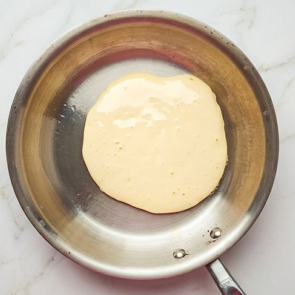 Une vue de dessus de pâte à crêpes beige pâle avec des bulles cuisant dans une poêle en acier inoxydable sur une surface de marbre blanc.