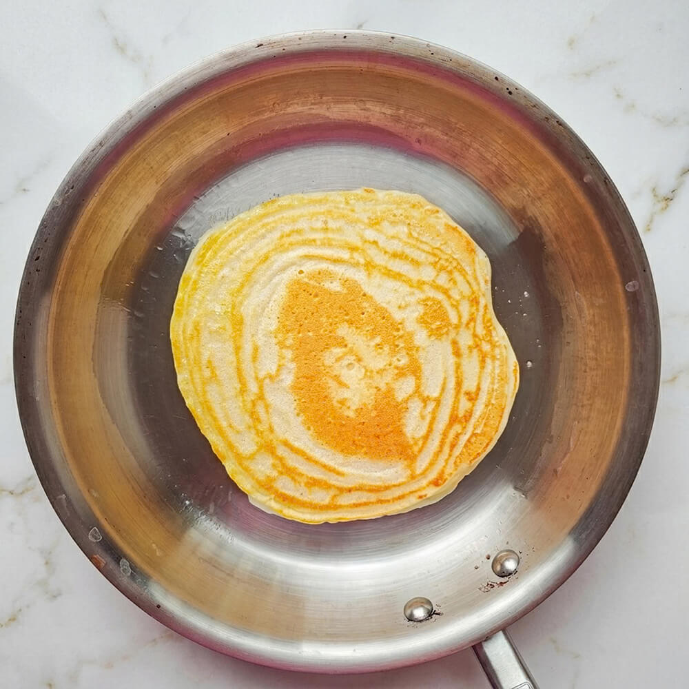 Une vue de dessus de pâte à crêpes beige dans un bol à motifs mouchetés marron avec une cuillère en acier inoxydable reposant dedans.