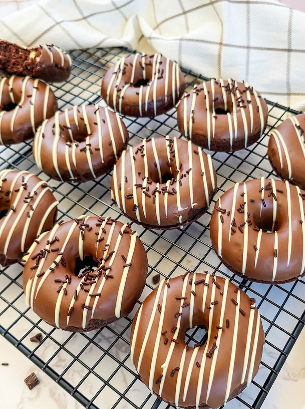 Des beignets au four, au chocolat disposés sur une grille de refroidissement noire avec une serviette à carreaux en arrière-plan. Chaque beignet est glacé d'une couche de glaçage au chocolat blanc et parsemé de vermicelles en chocolat.