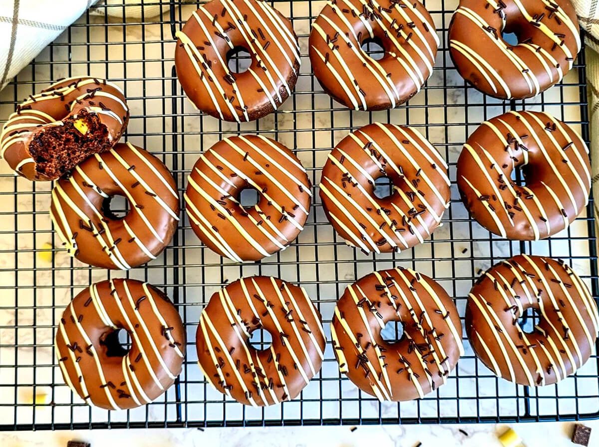 Des donuts au four chocolat cuits au four, disposés sur une grille de refroidissement noire, avec un glaçage brillant au chocolat au lait décoré de zébrures de chocolat blanc et de vermicelles. Un donut sur le côté est entamé pour révéler une texture moelleuse à l'intérieur.