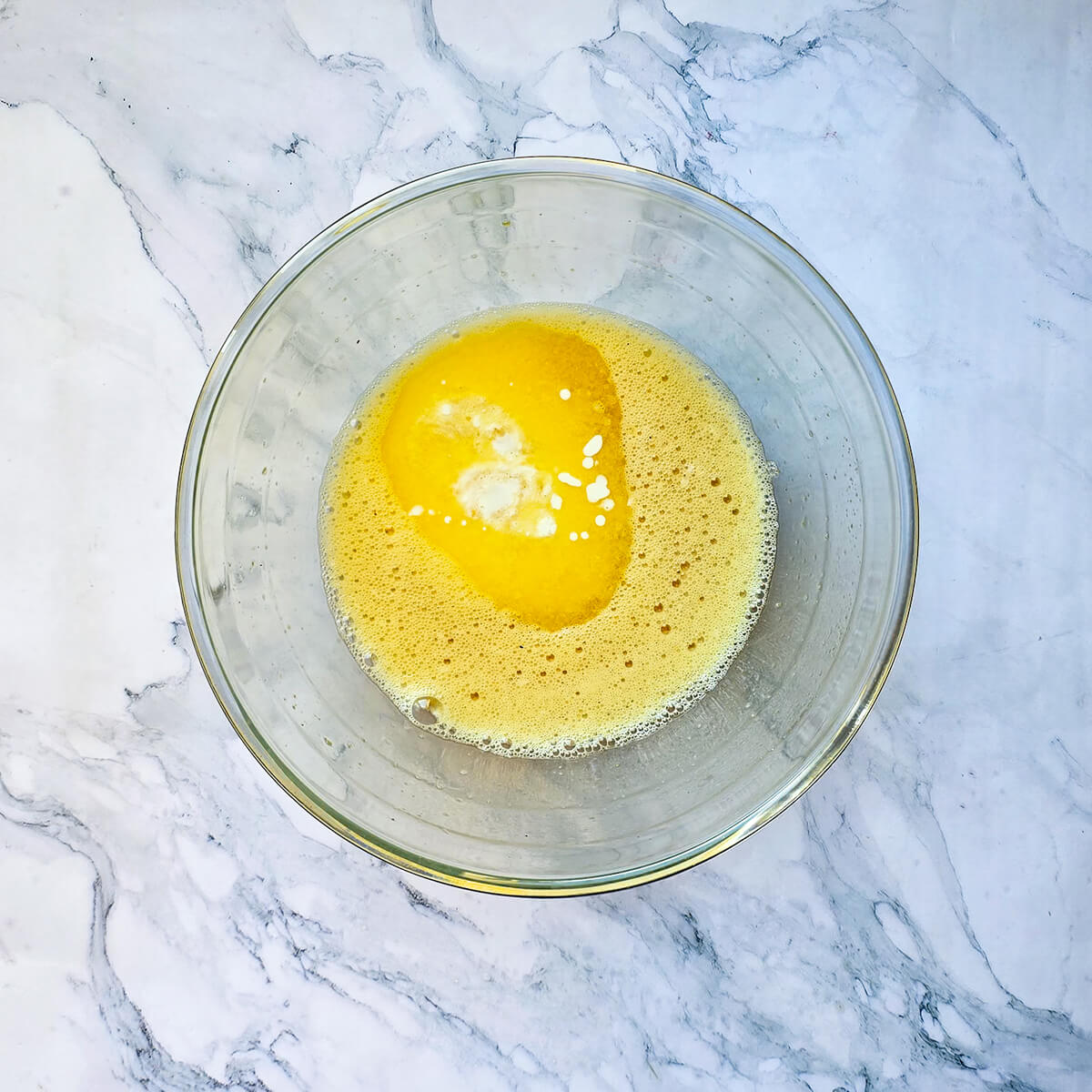 Un bol en verre contenant un mélange jaune pâle et mousseux d'œufs et de sucre battus, avec une petite flaque d'huile ou de beurre fondu au centre, sur un plan de travail en marbre.
