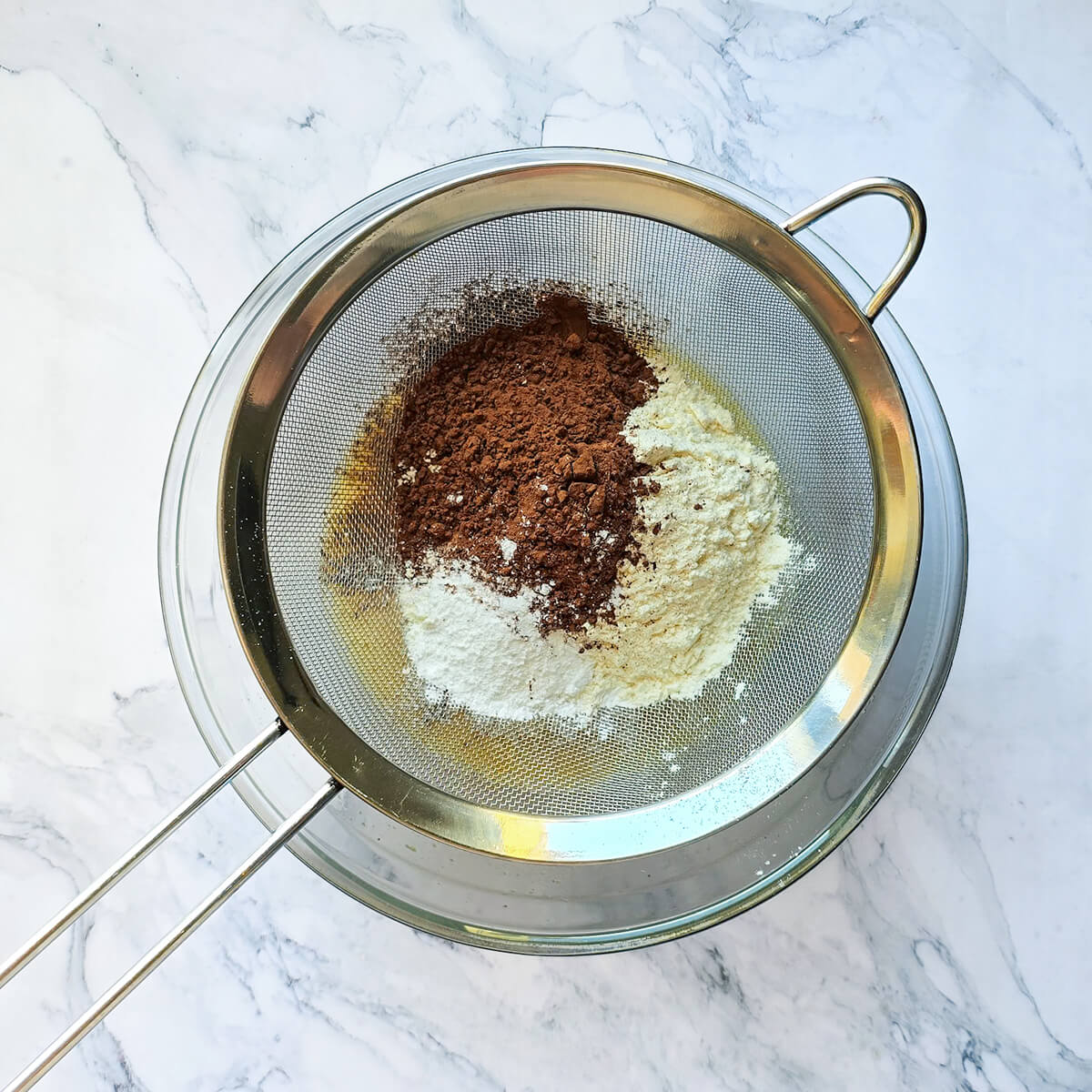 Vue de dessus d'un tamis à mailles fines en acier inoxydable posé sur un bol en verre. Le tamis contient une pile de cacao en poudre foncé et de farine blanche, prête à être tamisée.