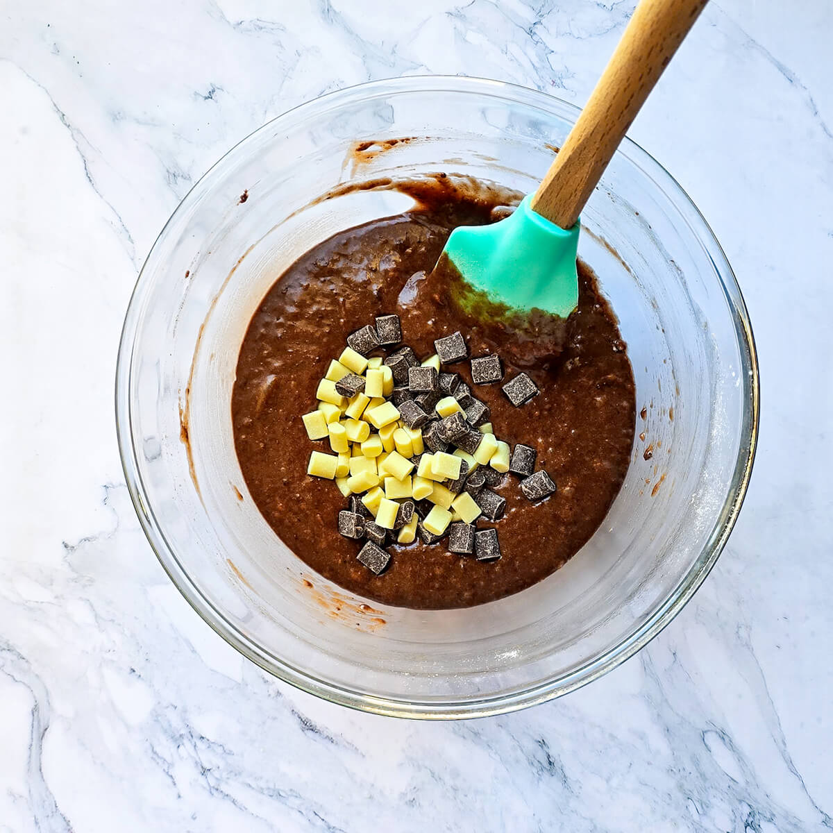 Un bol en verre contenant une pâte à donuts au chocolat lisse et brillante, avec un mélange de pépites de chocolat blanc et noir déposées au centre. Une spatule en silicone turquoise est partiellement immergée sur le côté.