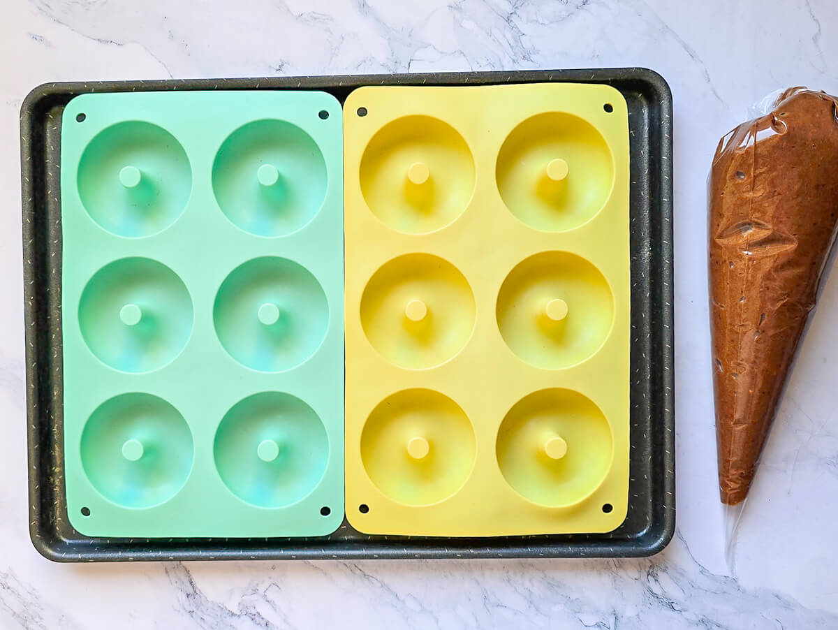 Deux moules à donuts en silicone de couleur vert et jaune et une poche à douille contenant la pâte à donuts, sur un fond en marbre.