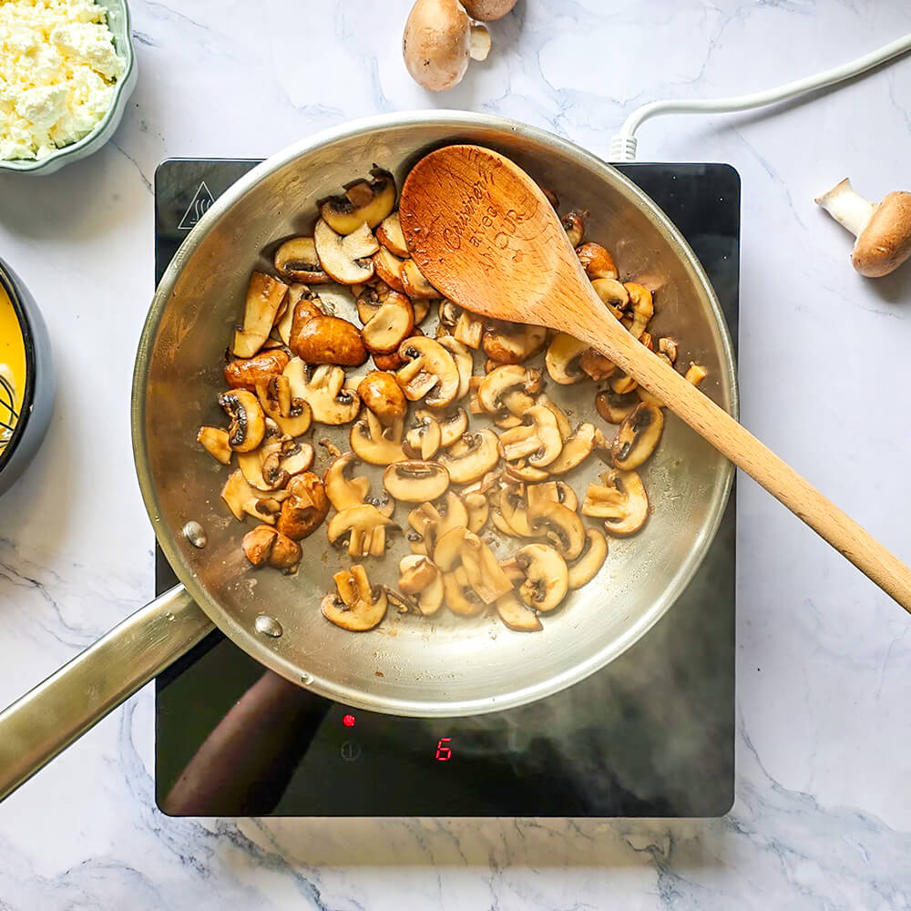 Une poêle en acier inoxydable sur une plaque à induction noire contient un œuf battu jaune vif en train de cuire. Des bols contenant des champignons cuits et de la feta émiettée sont visibles sur le plan de travail en marbre.