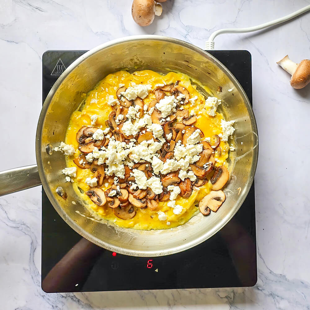 Une vue aérienne montre une poêle en acier inoxydable contenant une omelette aux œufs déjà cuite. Une généreuse garniture de champignons sautés et de feta émiettée est déposée sur un côté, prête à être pliée.