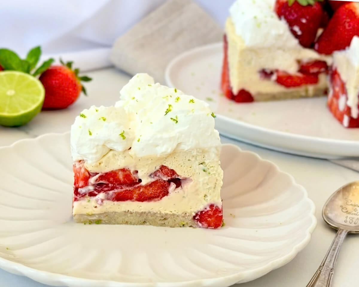 Une part de fraisier léger servie dans une assiette blanche ciselée, montrant les couches de biscuit, de crème onctueuse à la vanille et de morceaux de fraises fraîches, surmontée d'une chantilly aérienne et de zestes de citron vert.
