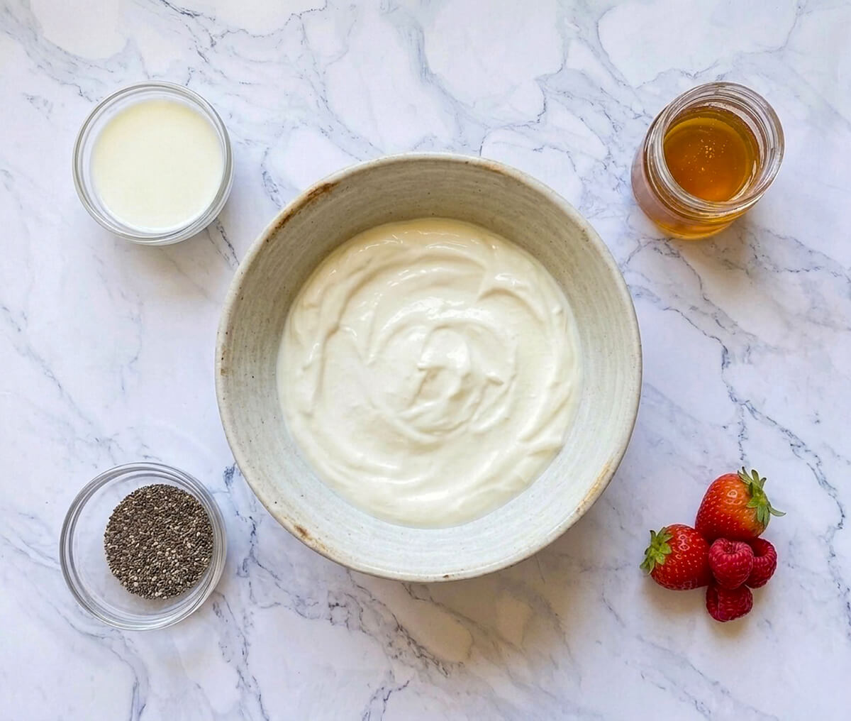 Vue de dessus à plat des ingrédients du pudding de chia sur un plan de travail en marbre blanc. Au centre, un grand bol de Skyr lisse entouré de quatre éléments : un petit bol de lait, un pot de miel doré, un ramequin de graines de chia noires et un petit tas de fraises et framboises fraîches.